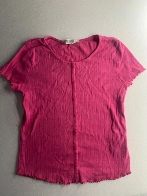 Rosa ribbad topp från H&M - Snygg rosa ribbad topp från H&M med korta ärmar och rund halsringning. Toppen har knappar framtill och en lätt vågig kant nertill för en extra detalj. Perfekt för dig som gillar färg och enkel stil.💞