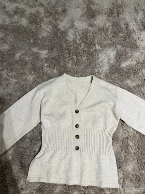 Beige ribbad kofta med knappar - Säljer en beige kofta med ribbad midja och fyra bruna knappar framtill. Koftan har v-ringning och långa ärmar. Materialet är mjukt och stretchigt, vilket gör den bekväm att bära. Obs: kommer från shein 