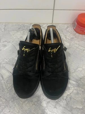 Svarta sneakers från Giuseppe Zanotti - Svarta sneakers med guldfärgade dragkedjor och signaturdetalj på plösen. Skorna har en låg siluett, svarta skosnören och ovandel i mocka och läder. Insidan är beige och sulan är svart. Perfekta för dig som gillar exklusiv och stilren streetwear.