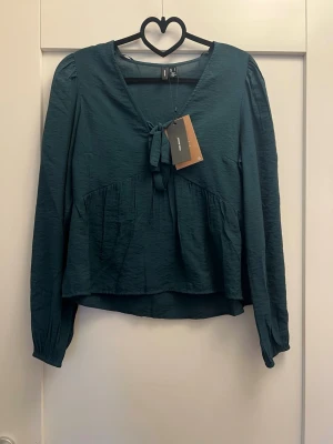 Mörkgrön blus med knyt från Vero Moda - Supersnygg mörkgrön blus från Vero Moda med långa ärmar och v-ringning med knyt framtill. Blusen har lätt rynkade axlar och en lös, flowy passform. Materialet är tunt och har en lätt struktur som ger en cool look.