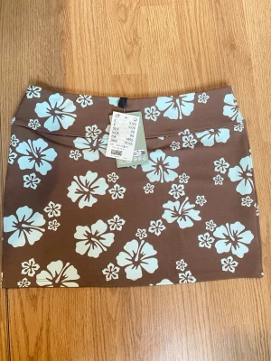 Brun blommig minikjol från Divided - Säljer en brun mini kjol från Divided med ljus blåa blommor. Kjolen har en tight passform och är tillverkad i mjuk polyester med stretch. Perfekt för dig som vill ha en trendig och somrig look!