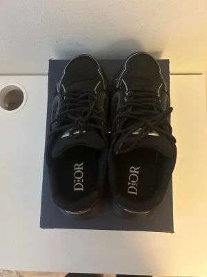 Svarta Dior sneakers med silvriga detaljer - Snygga svarta sneakers från Dior med silvriga detaljer och logga på hälen. Skorna har en chunky sula och vita kontrastsömmar som ger en modern look. Ovandelen är i textil och syntet, med klassisk snörning och bekväm passform.