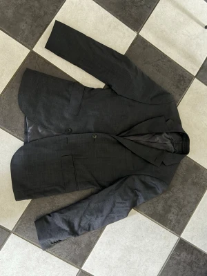 Mörkgrå kavaj från Riley - Snygg mörkgrå kavaj från Riley med klassisk passform och två knappar framtill. Kavajen har diskreta fickor och är helfodrad med slät insida. Perfekt för en stilren look och passar till många olika outfits. Passar S/M. Använt 1 gång.