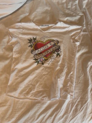 Vit t-shirt Zadig & Voltaire hjärta - Vit t-shirt från Zadig & Voltaire med coolt tryck av ett hjärta och blommor på bröstet. T-shirten har korta ärmar och rund halsringning med råa kanter. 