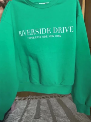 Grön sweatshirt Riverside Drive - Snygg grön sweatshirt med texten 'RIVERSIDE DRIVE UPPER EAST SIDE, NEW YORK' tryckt i vitt på bröstet. Tröjan har rund halsringning, ribbade muddar och är gjord i mjukt bomullsmaterial. Perfekt för en avslappnad streetwear-look.