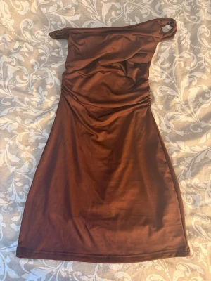 Brun silkes klänning - Snygg brun klänning i stretchigt material med silkes material. Klänningen är aldrig använd och har inga hål eller något.  Klänningen har en åtsittande passform och är kort i modellen, perfekt för dig som vill sticka ut. Tyget har en lätt glans och fina draperingar vid överdelen.