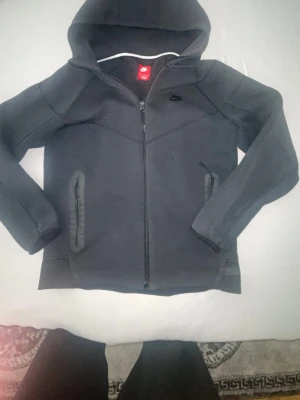 Svart Nike hoodie med dragkedja - Snygg svart hoodie från Nike med hel dragkedja framtill och två fickor med dragkedja. Klassisk huva och diskret Nike-logga på bröstet. Perfekt för en sportig och avslappnad stil. Tillverkad i mjukt material för extra komfort.