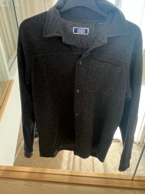Mörkgrå skjorta från Jack & Jones - Snygg mörkgrå skjorta från Jack & Jones med klassisk krage, bröstficka och knappar framtill. Skjortan har lång ärm och är tillverkad i ett mjukt material som känns bekvämt mot huden. Perfekt för en avslappnad men stilren look.