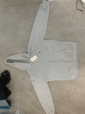Grå zip-hoodie från Lacoste - Snygg grå hoodie med dragkedja från Lacoste. Klassisk krokodillogga på bröstet, två stora fickor framtill och skön huva. Perfekt för en avslappnad stil. Tillverkad i mjukt bomullsmaterial. Köpte nyss kunde inte lämna eftersom blev försenad 