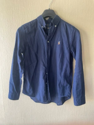 Marinblå skjorta från Ralph Lauren - Klassisk marinblå linne skjorta från Ralph Lauren. Skjortan har långa ärmar, knappar framtill och är tillverkad linne. Perfekt till en avslappnad men ändå stilren look.