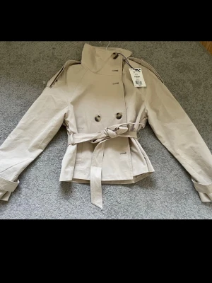 Berskha trenchcoat  - Stilren beige trenchcoat från Berskha med dubbelknäppning, höga axelklaffar och knytskärp i midjan. Jackan har en kortare modell och hög krage, perfekt för en trendig look. Tillverkad i ett slätt bomullsliknande material.