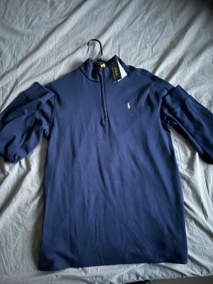 Marinblå halvzip från Polo Ralph Lauren - Snygg marinblå långärmad tröja från Polo Ralph Lauren med halv dragkedja och klassisk broderad logga på bröstet. Perfekt för en stilren look och enkel att matcha med jeans eller chinos.