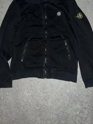 Svart hoodie från Stone Island. PRIS KAN DISKUTERAS! - Svart hoodie från Stone Island med dragkedja framtill, två snedställda fickor med dragkedja och klassisk kompasspatch på vänster ärm. Mjuk bomullskänsla och huva för extra stilpoäng. Perfekt för dig som gillar streetwear och snygga detaljer.
