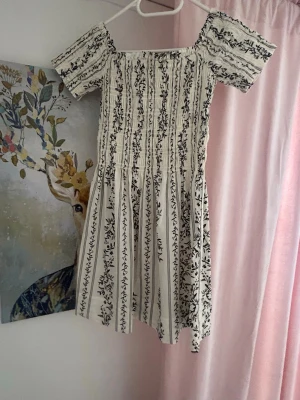 Vit kortklänning med blommigt mönster - Supersöt vit kortklänning med svarta blommiga ränder och smockad överdel. Klänningen har korta puffärmar och en luftig kjol, perfekt för varma dagar. ☀️✨