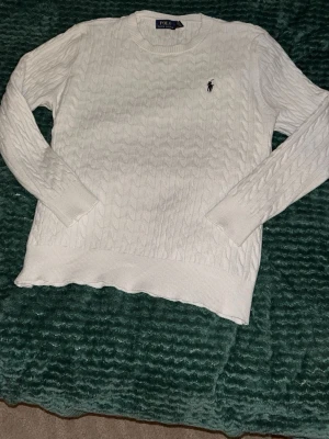 Vit kabelstickad tröja Polo Ralph Lauren - Kabelstickad vit tröja från Polo Ralph Lauren med rund halsringning och den klassiska broderade loggan på bröstet. Tröjan har långa ärmar och ribbade muddar vid ärmslut och nederkant. Perfekt för en clean och stilren look.