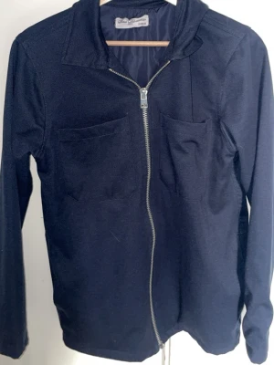 Mörkblå overshirt med dragkedja - Stilren mörkblå overshirt med två bröstfickor och hel dragkedja framtill. Jackan har klassisk krage och är tillverkad i ett mjukt tyg som känns bekvämt. Perfekt lager-på-lager-plagg för en clean look. Och PERFEKT nu inför sommaren! XS på tröjan men passar mig bra och jag är 181. 