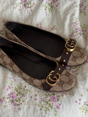 Bruna ballerinaskor  - Snygga ballerinaskor som jag köpt från Vinted i beige och brunt. Skorna har rund tå, mörkbrunt läderband med dubbla guldfärgade spännen framtill. Lite småskavanker här o där men inget som syns när man har på sig dom. Innermåttet är ca 24 cm om man mäter från mitten av hålen till toppen. 