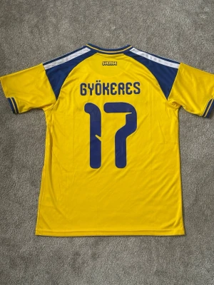 Sverige fotbollströja Gyökeres 17 - Gul och blå Sverige fotbollströja från Adidas med Gyökeres och nummer 17 på ryggen. Tröjan har korta ärmar, rund hals och broderat landslagsmärke på bröstet. Klassisk design med blå detaljer och vita ränder på axlarna. Perfekt för fotbollsfans! 26/27 vm tröjan. Ny aldrig använd