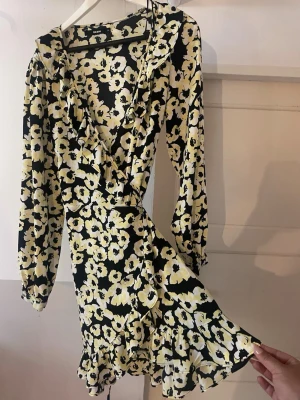 Blommig omlottklänning från BikBok - Supersnygg kort omlottklänning från BikBok med svart botten och stora gula och vita blommor. Klänningen har volangdetaljer, v-ringning och långa ärmar. Perfekt för dig som vill sticka ut med ett färgglatt mönster och skön passform.