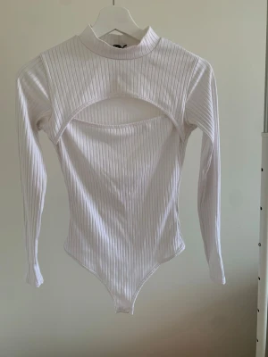 Vit ribbad långärmad body - Säljer en vit ribbad body med lång ärm och hög hals. Bodyn har ett coolt cut-out parti framtill som ger en trendig look. Materialet är stretchigt och bekvämt, perfekt för att styla med jeans eller kjol. Passar dig som gillar minimalistisk och modern design.