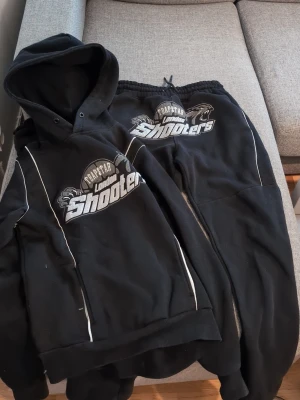 Trapstar London Shooters Tracksuit svart - Svart hoodie från Trapstar med stor 'London Shooters'-logga framtill i vitt och grått. Hoodien har huva, vita kontrastsömmar och en sportig vibe. Perfekt för dig som gillar streetwear och vill sticka ut med en fet logga.
