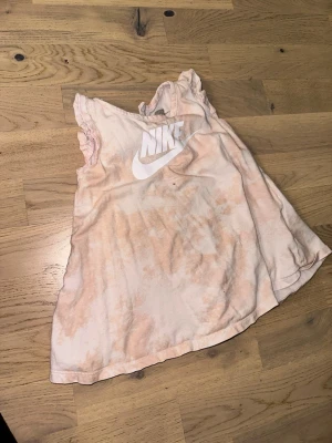 Ljusrosa ärmlös topp från Nike - Ljusrosa ärmlös topp från Nike Nike topp i ljusrosa med vitt logotryck framtill. Modellen är ärmlös med volangdetaljer vid axlarna och har en avslappnad passform. Tillverkad i mjuk bomull, perfekt för en sportig och chill stil.