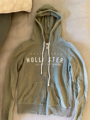 Grön hoodie från Hollister  - Snygg grön hoodie från Hollister med dragkedja och vit text på bröstet. Modellen har huva med vita snören och två fickor framtill. 