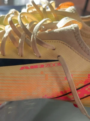 Nike Air Zoom gula fotbollsskor - Säljer ett par gula Nike Air Zoom fotbollsskor med svart swoosh och snörning. Skorna har en lätt och smidig design i syntetmaterial, perfekta för snabba rörelser på planen. Snygg grafik i orange och rosa detaljer på sidan. Grymma skor för dig som vill sticka ut!