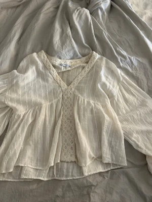 Fin blus Zara - Luftig vit blus från Zara med v-ringning och dekorativa spetsdetaljer framtill. Blusen har ett lätt transparent tyg med diskreta ränder och vida, långa ärmar. Perfekt för en avslappnad och trendig look.