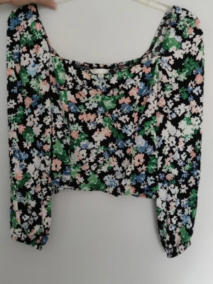 Blommig blus med puffärm - Blus med croppad passform och långa puffärmar. Mönstret är i svart, vit, grön, blå och rosa. Blusen har smockad rygg och lätt v-ringad hals..
