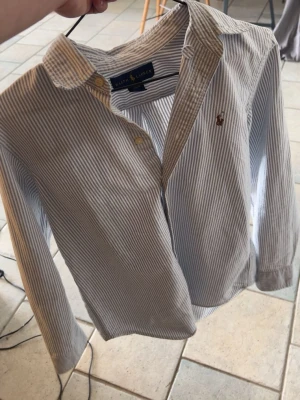 Randig skjorta från Ralph Lauren - Klassisk skjorta från Ralph Lauren med smala blå och vita ränder, broderad logga på bröstet och knappar framtill. Skjortan har lång ärm och krage, perfekt för en stilren look. Passar 170cm