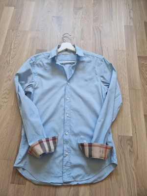  Burberry skjorta - Snygg ljusblå skjorta från Burberry med klassiska rutiga detaljer på insidan av manschetterna och broderad logga på bröstet. Skjortan har lång ärm, knappar framtill och är tillverkad i bomull. Perfekt för en stilren look.