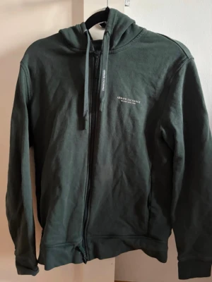 Mörkgrön hoodie från Armani Exchange - Snygg mörkgrön hoodie med dragkedja från Armani Exchange. Klassisk passform med huva och dragsnören, samt diskret logga på bröstet. Tillverkad i mjukt bomullsmaterial som känns skönt mot huden. Perfekt för en avslappnad och trendig look.