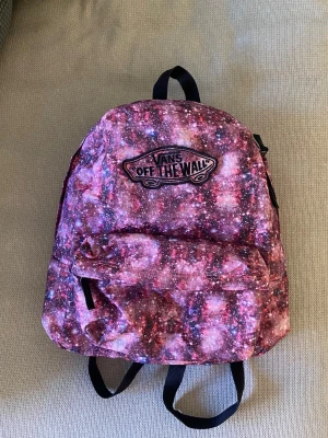 Vans ryggsäck med galaxmönster - Ryggsäck från Vans med ett unikt galaxmönster i rosa, lila och svart. Ryggsäcken har ett stort huvudfack, en mindre ficka framtill och justerbara svarta axelremmar. 