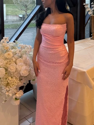 Ljusrosa axelbandslös långklänning - Elegant ljusrosa axelbandslös långklänning med slits på sidan och markerad midja. Korsett i bak som går att ljustera med band. Klänningen är skräddarsydd i Kosovo. Storleken skulle jag säga är mellan 36-38