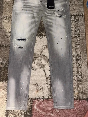 Grå slitna jeans från Dsquared2 - Säljer ett par grå jeans från Dsquared2 med skinny fit, slitningar och hål på framsidan samt färgstänk i vitt och blått. Jeansen har en tvättad look och klassisk femficksmodell. Perfekta för dig som gillar streetstyle och trendiga detaljer.