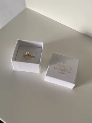 Guld ring pärla Caroline Svedbom - Snygg guldfärgad ring med pärla från Caroline Svedbom.  Lite sliten inuti men inget som märks när man har den på.  Säljer då den är för stor för mig Gjord i mässing med 18 karats guldplatering Nypris 395kr