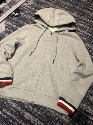 Grå Moncler hoodie med dragkedja - Snygg grå hoodie från Moncler med dragkedja framtill och huva med dragsko. Ärmsluten har ränder i rött, vitt och blått för en sportig look. Moncler-logga på ena ärmen. Mjuk bomullsblandning som är skön att ha på sig.