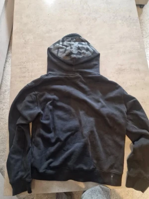 Svart syna world hoodie - Svart hoodie med känguruficka och snörning i huvan. Huvan har en snygg camo-detalj på insidan som ger en cool kontrast. Perfekt för en avslappnad streetstyle-look. Mjuk bomullskänsla och ribbade muddar vid ärmslut och midja.