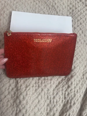 Röd glittrig kuvertväska Marc Jacobs - Snygg röd kuvertväska från Marc Jacobs med glittrig finish och guldfärgad dragkedja. Väskan har en stilren rektangulär form och logga i guld på framsidan. Perfekt accessoar för att lyfta din outfit med lite extra bling.