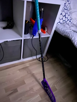 Innebandyklubba svart/lila - Säljer en innebandyklubba med svart skaft och lila blad. Skaftet har ett mönster i regnbågsfärger och greppet är turkost. Perfekt för dig som gillar att spela innebandy och vill ha en färgglad klubba.