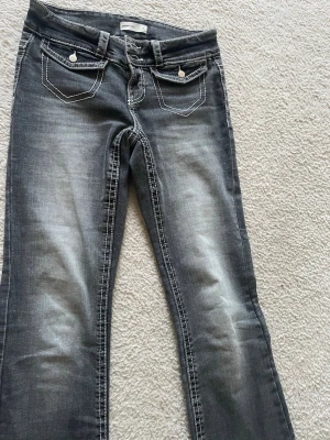 Grå bootcut jeans med vita sömmar - Snygga grå bootcut jeans från Gina Tricot med markerade vita sömmar och dubbla knappar i midjan. Jeansen har två fickor fram med lock och dekorativa knappar samt slitningar för en cool look. Perfekta för dig som gillar en avslappnad men trendig stil.