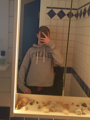 Grå hoodie från GANT med tryck - Snygg grå hoodie från GANT med klassiskt tryck på bröstet och dragsko i huvan. Mjuk bomullskänsla och avslappnad passform, perfekt till jeans eller mjukisbyxor. Huvan har vita snören och tröjan har ribbade muddar nertill.