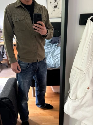 Olivgrön overshirt från Nimbus CPH - Olivgrön overshirt från Nimbus CPH med två bröstfickor och knäppning framtill. Tillverkad i ett slitstarkt bomullstyg med klassisk krage och raka ärmar. Perfekt att bära öppen eller stängd för en avslappnad stil. 