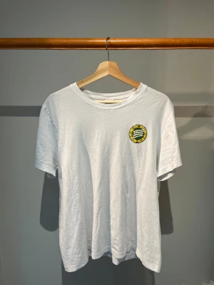 HAMMARBY SÖDERMALM - Vit t-shirt med rund hals och korta ärmar. På bröstet finns Hammarbys klubbmärke i grönt och gult, och på ryggen står det 'HAMMARBY SÖDERMALM' i stora gröna bokstäver. Perfekt för dig som vill visa ditt stöd för Hammarby.