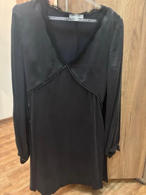 Svart långärmad blus med spetsdetalj - Stilren svart blus med långa ärmar och vacker spetsdetalj längs ringningen. Blusen har en mjuk, något glansig yta som ger en elegant känsla. Perfekt för dig som gillar subtila detaljer och en klassisk look.