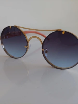 Runda solglasögon med guldiga detaljer - Snygga runda solglasögon med guldiga metallbågar och blåtonade glas. Skalmarna är orange med dekorativa gulddetaljer. Perfekt accessoar för att sticka ut och ge din stil en cool touch.