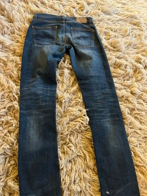 Blå smala jeans från Nudie Jeans - Säljer ett par blå smala jeans från Nudie Jeans med klassisk femficksmodell och snygga slitningar. Jeansen har kontrastsömmar och patch bak i midjan. Tillverkade i denim med en cool tvätt som ger en avslappnad look.