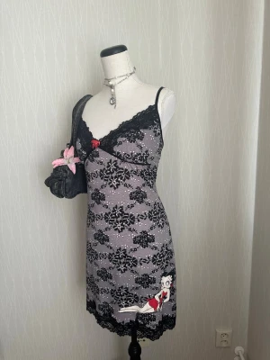 Grått nattlinne med Betty Boop-tryck - Supersött grått nattlinne med svarta spetsdetaljer och mönster, samt en röd rosett framtill. Nedtill finns ett tryck av Betty Boop i röd klänning. Smala axelband och figurnära passform. Perfekt för dig som gillar en lekfull och trendig stil.