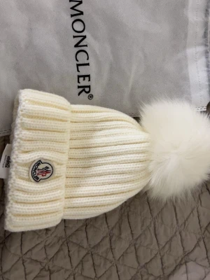 Vit riktig Moncler mössa med pälstofs - Pris kan självklart diskuteras. Helt ny moncler oanvänd snygg och lyxig vit stickad mössa från Moncler med bred uppvikt kant och stor fluffig pälstofs på toppen. Mössan har Monclers klassiska logga framtill och är tillverkad i mjuk ull. Perfekt accessoar för kalla dagar och en stilren vinterlook. 